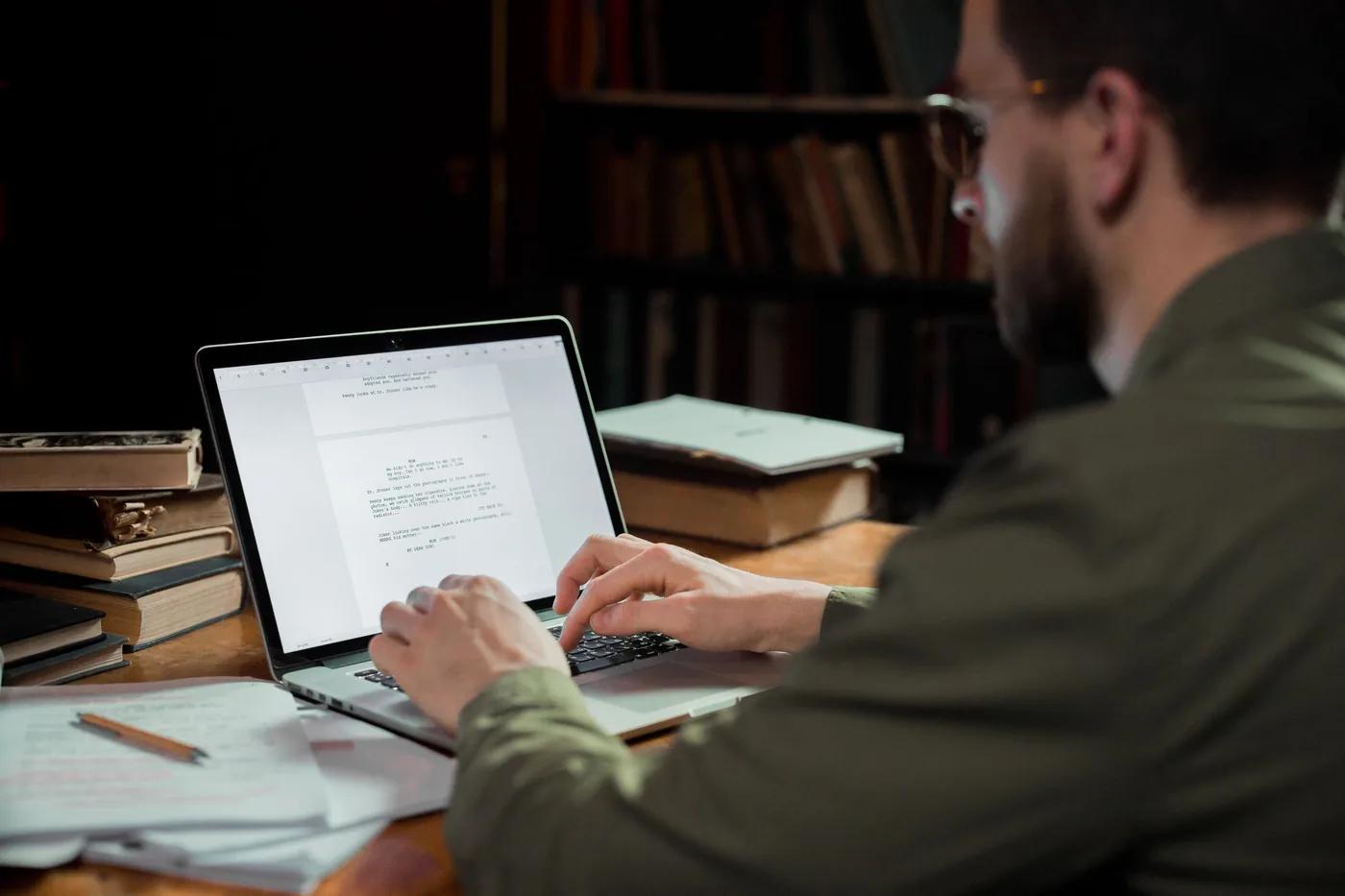 Someone typing a document on a laptop at a desk surrounded by stacked books and handwritten notes