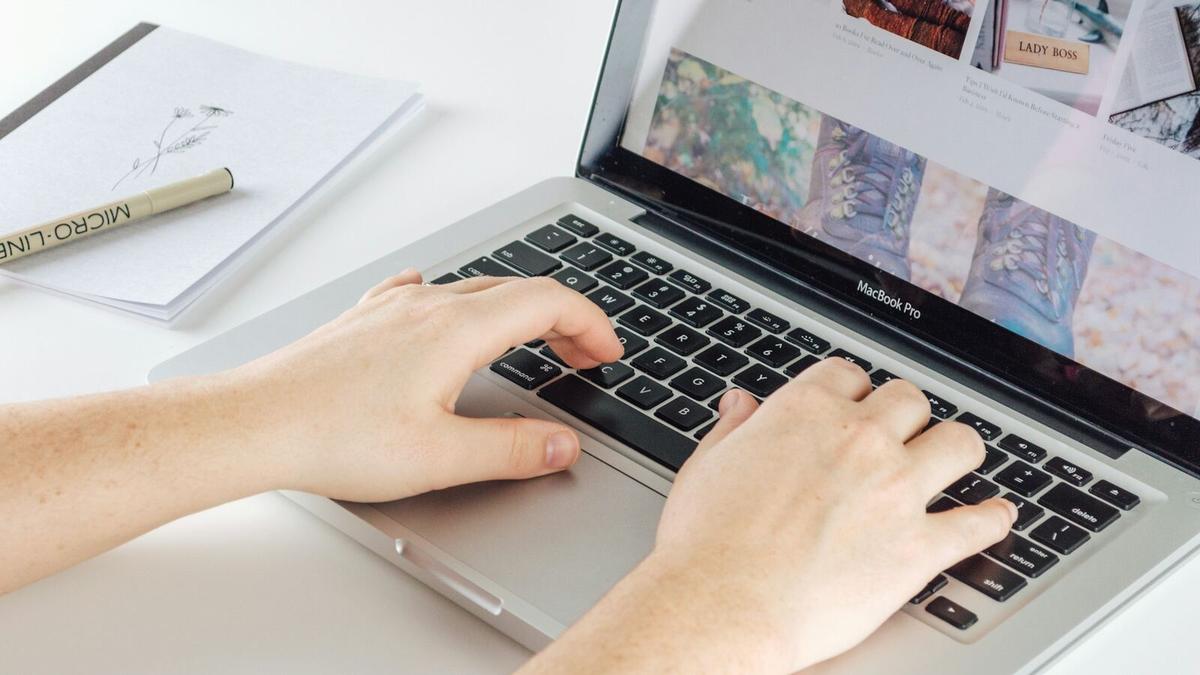 Hands typing on a laptop at a white desk, a notebook and pen beside it