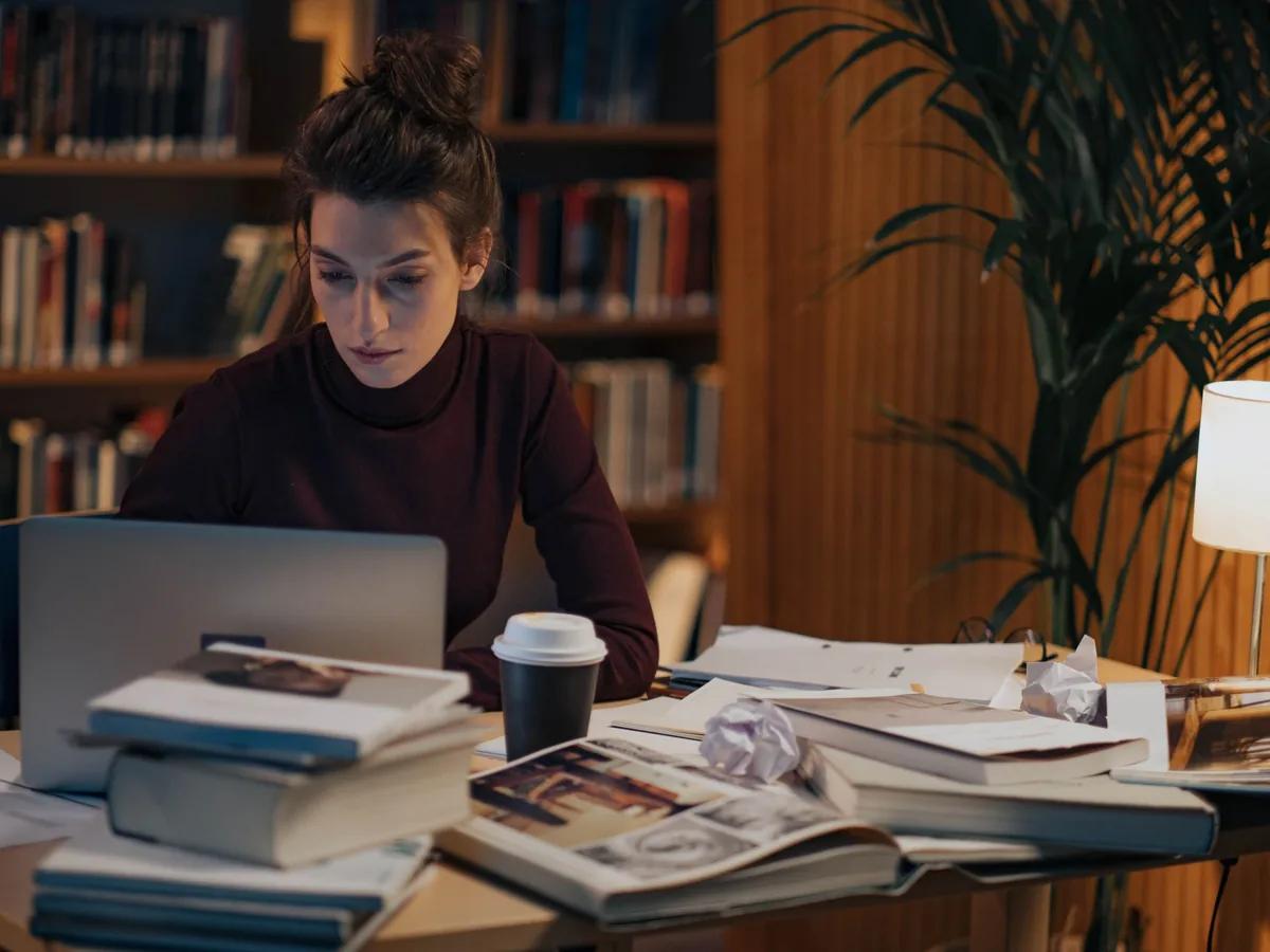 Someone working at a laptop at a library desk stacked with open books and papers