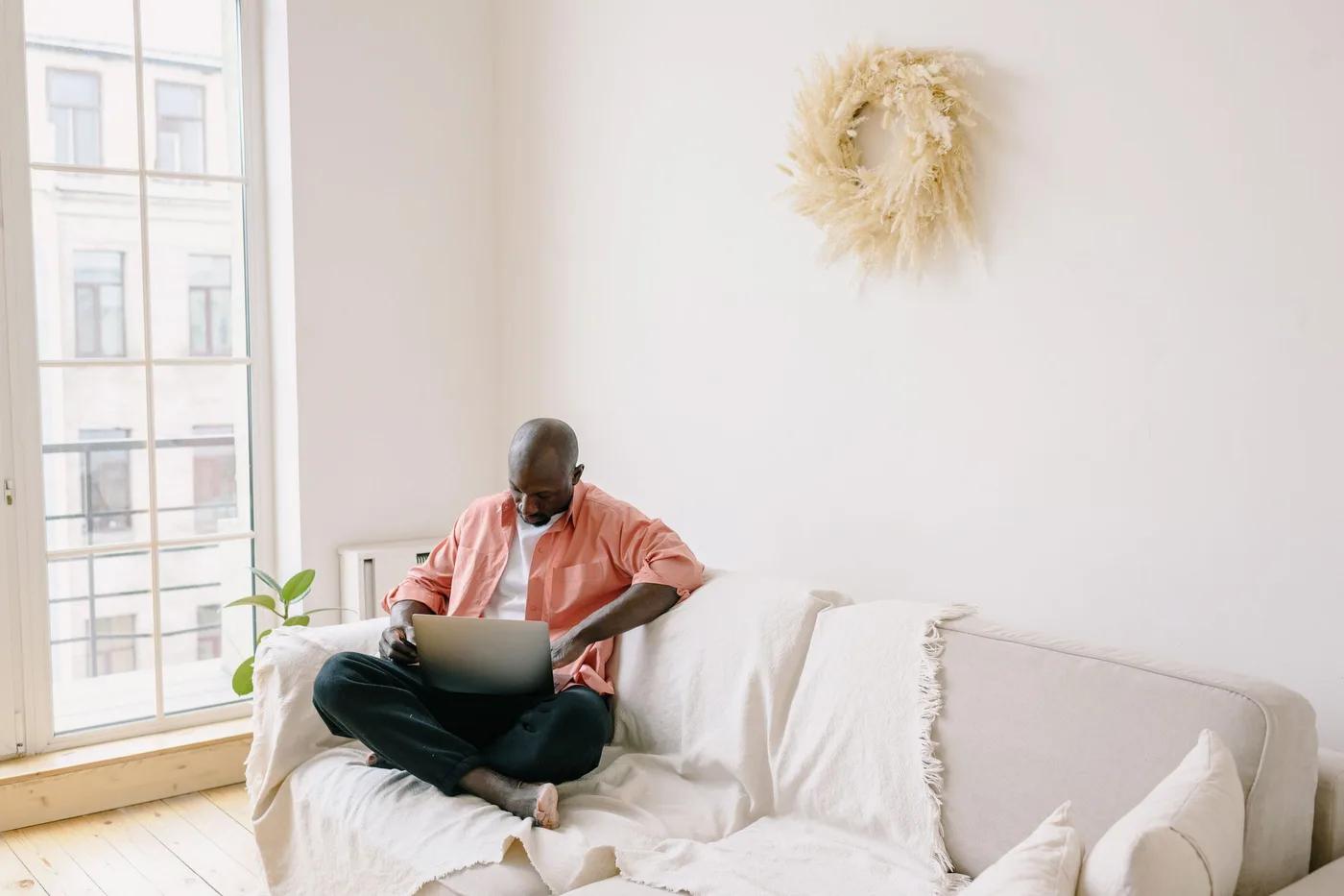 Someone sitting cross-legged on a sofa with a laptop in a bright room beside a tall window